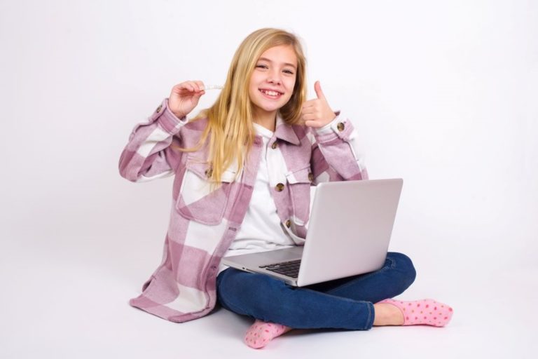 A young girl gives a thumbs up holding an Invisalign aligner and working on a laptop.